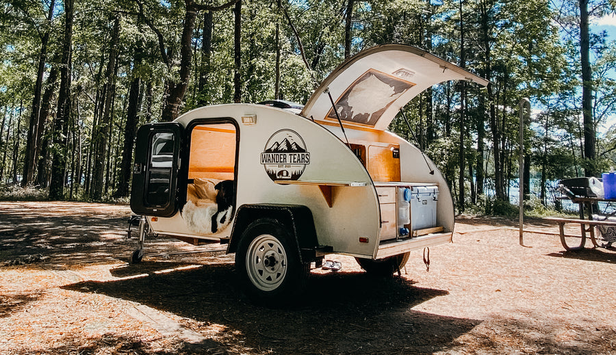 A compact white teardrop camper (an ideal RV rental) with its dog companion at a sunny forest campsite by a lake.