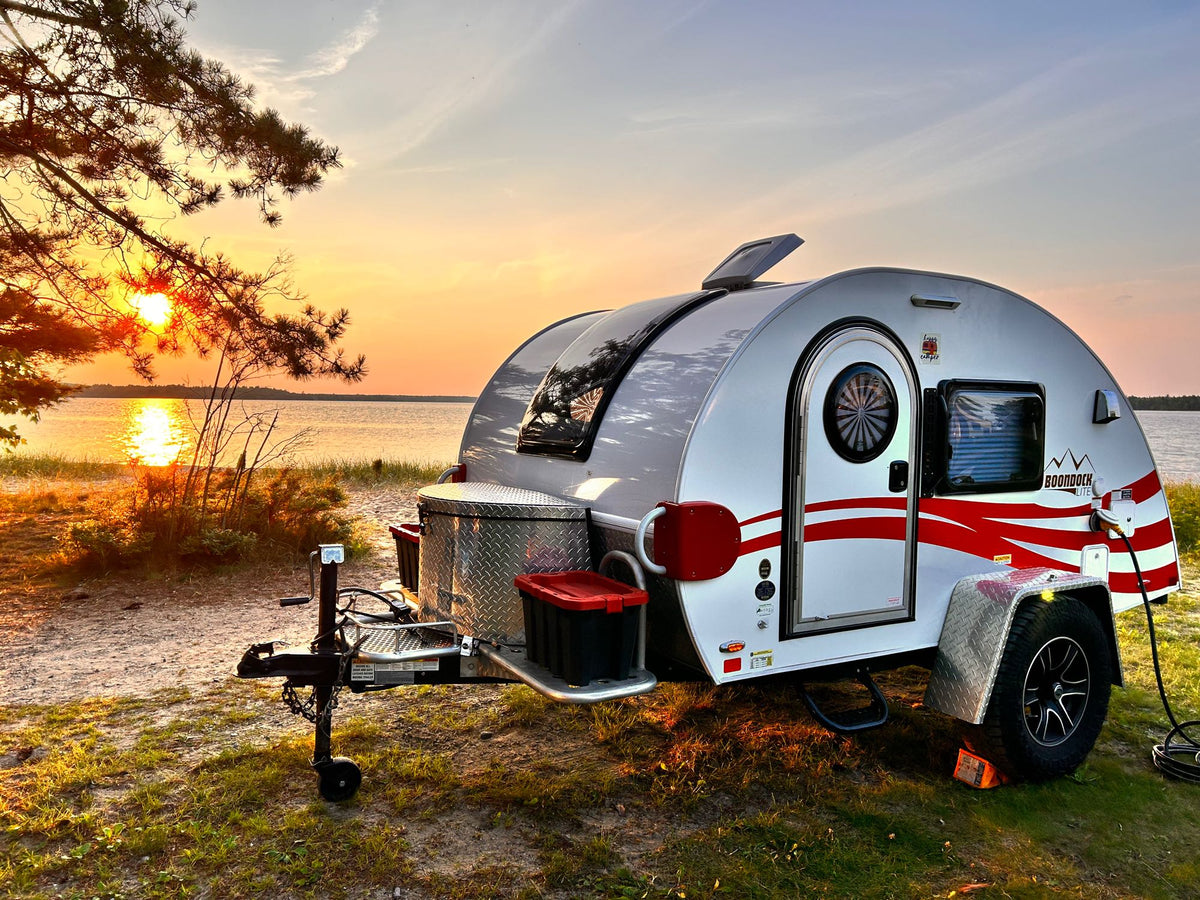 Enjoy an RV rental adventure! A white teardrop camper with red accents sits by a lake at sunset