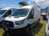 Front view of a white Ford Transit van, set up as an RV rental, with a roof cargo box and ladder rack. Parked in a sunny outdoor lot.