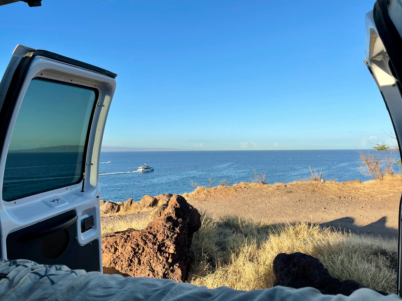 Scenic ocean view from an RV rental: a boat glides by a rocky coastline, framed by the open rear of a camper van.