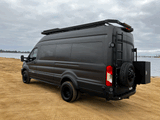 A sleek, dark grey Ford Transit adventure camper van with custom roof rack and rear ladder, ideal for RV rental, parked by water.