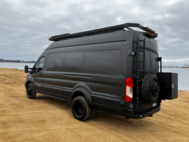 A sleek, dark grey Ford Transit adventure camper van with custom roof rack and rear ladder, ideal for RV rental, parked by water. — photo 9