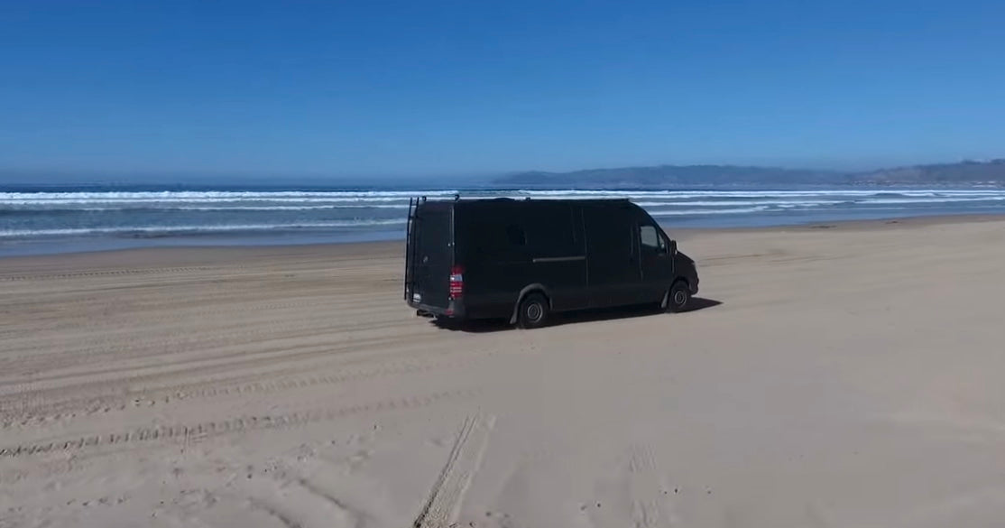 Black camper van parked on a sunny sandy beach with ocean waves. Ideal rv rental for coastal adventures.