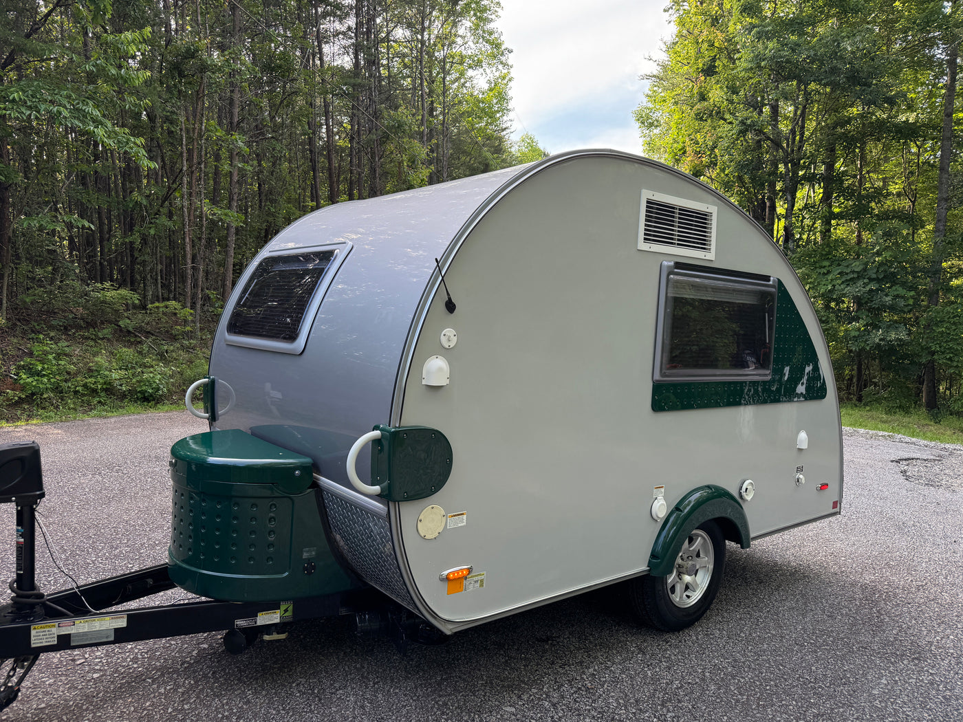 Grey & green teardrop RV rental parked on an asphalt road in a green forest, ready for travel. — photo 3