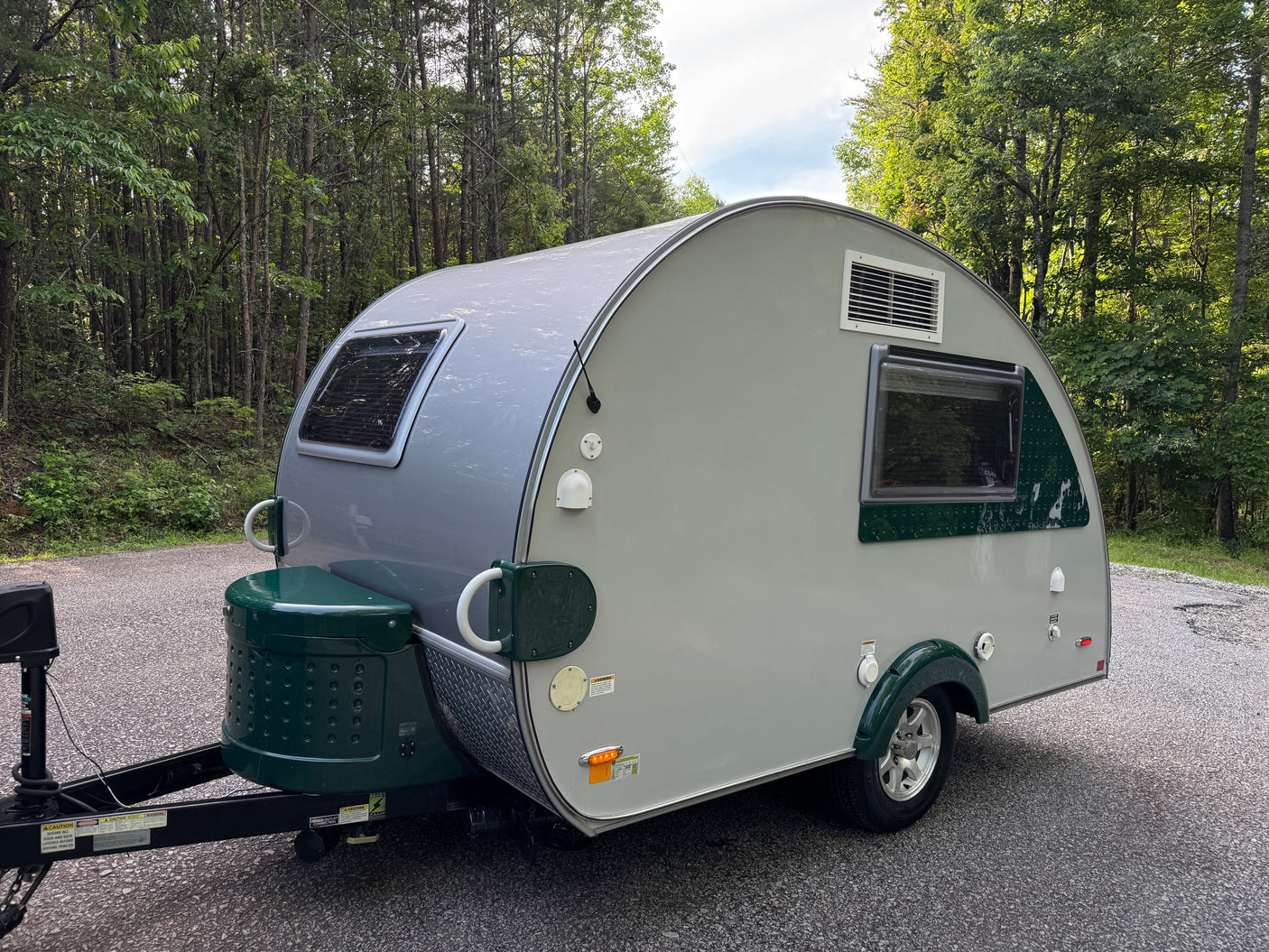 Grey & green teardrop RV rental parked on an asphalt road in a green forest, ready for travel.
