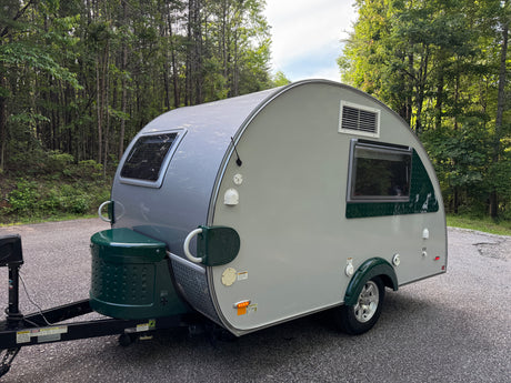 Grey & green teardrop RV rental parked on an asphalt road in a green forest, ready for travel.