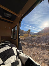 Cozy RV rental with a checkered blanket open to a stunning desert mountain landscape. Sun shines on a Yucca in the rocky terrain.