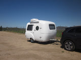 A white Happier Camper travel trailer, ideal for RV rental, hitched to an SUV in a sunny vineyard setting.