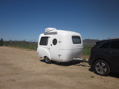 A white Happier Camper travel trailer, ideal for RV rental, hitched to an SUV in a sunny vineyard setting.