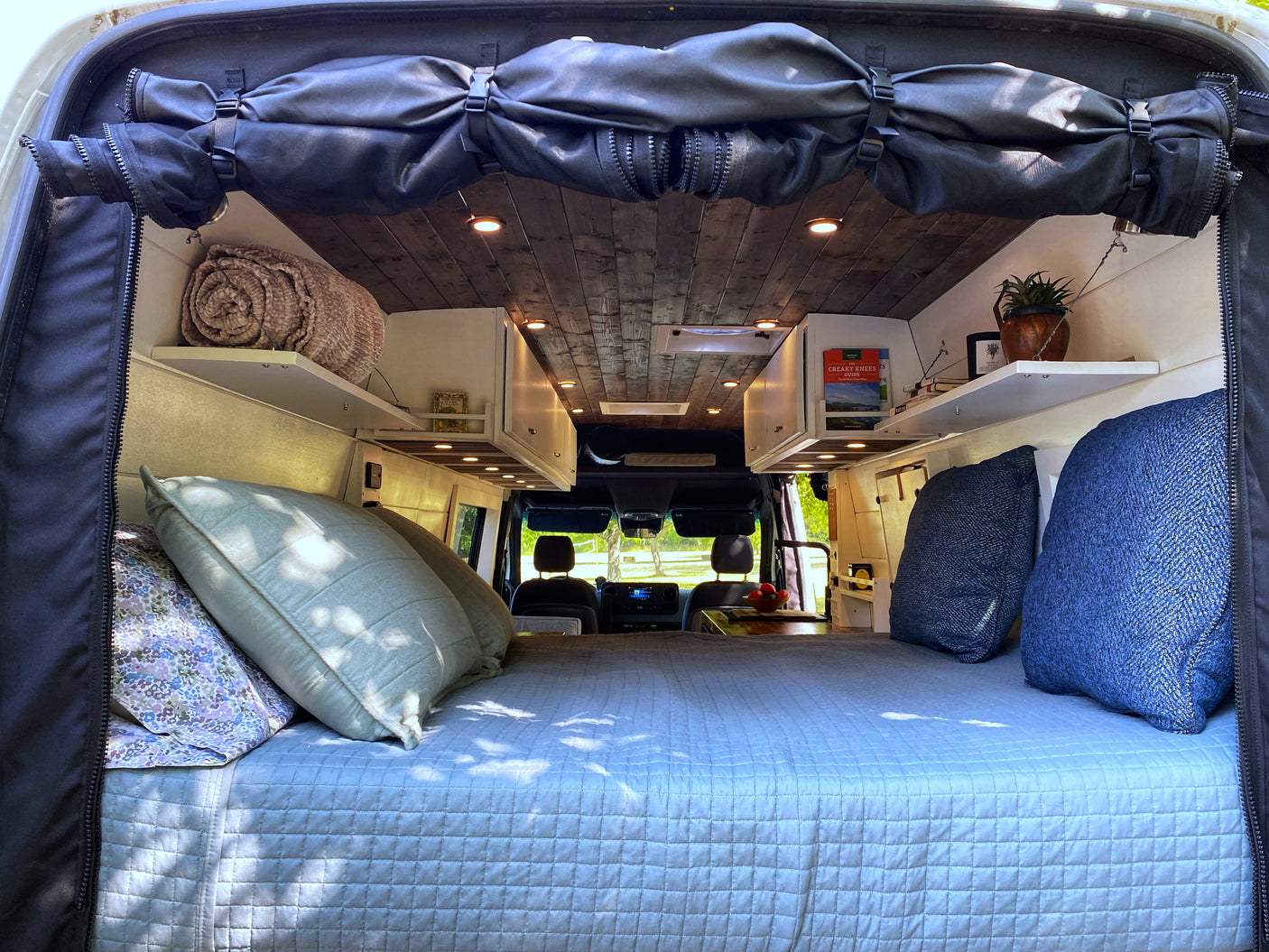 Cozy RV rental interior featuring a large bed, overhead storage, and wooden ceiling. View from the rear to the front cab.