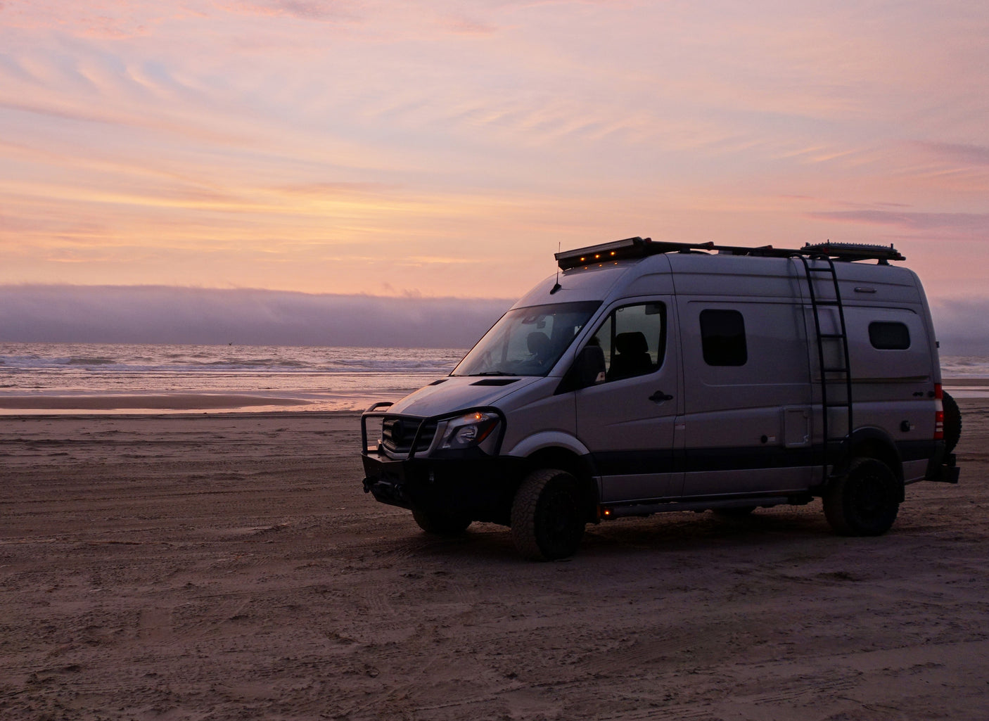A custom RV rental camper van parked on a sandy beach at sunset, with a misty ocean horizon behind.