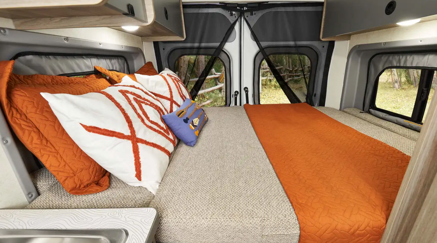 RV rental interior: a comfy bed with orange bedding, overhead storage, and mesh screens revealing peaceful forest views.
