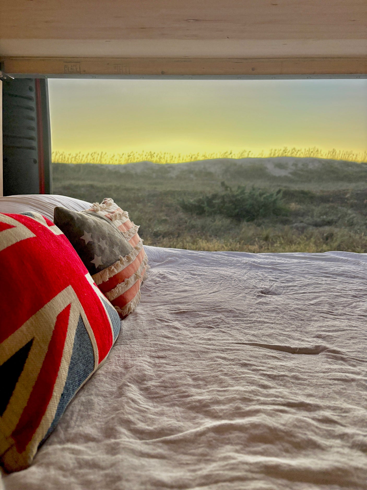RV rental bed with patriotic pillows (Union Jack, US flag) offering a serene view of dunes under a soft yellow sky.