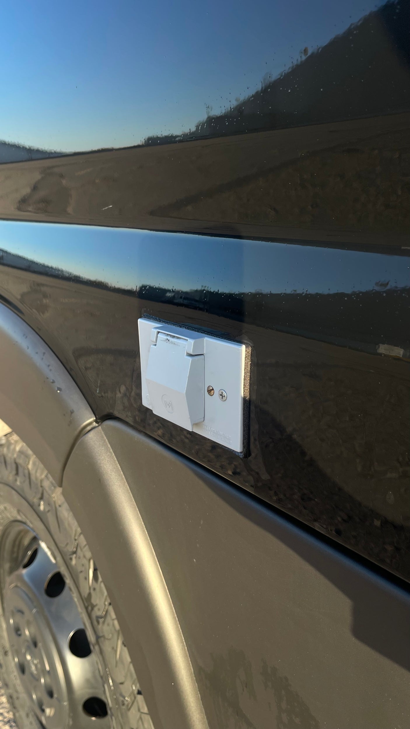Light gray exterior power outlet on an RV rental van, ready for shore power connection. The vehicle's black paint reflects the sky.