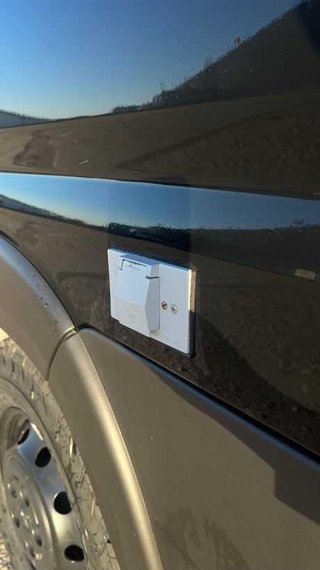 Light gray exterior power outlet on an RV rental van, ready for shore power connection. The vehicle's black paint reflects the sky.