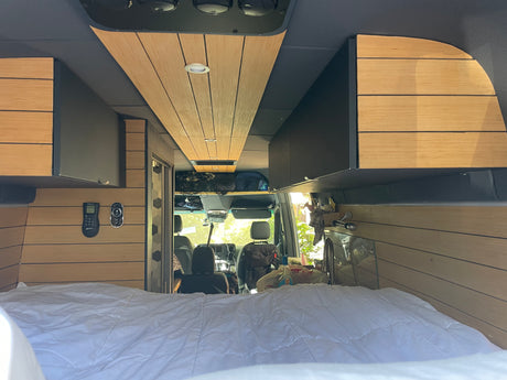 View from the bed of a spacious RV rental, featuring sleek wood paneling, storage cabinets, and front cockpit.