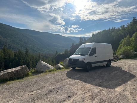 White RV rental van at a scenic mountain overlook. Lush green forests fill the valley under a bright, cloudy sky.
