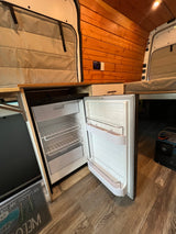 Open refrigerator in an rv rental van, showing wood paneling, controls, and a portable power station on a laminate floor.