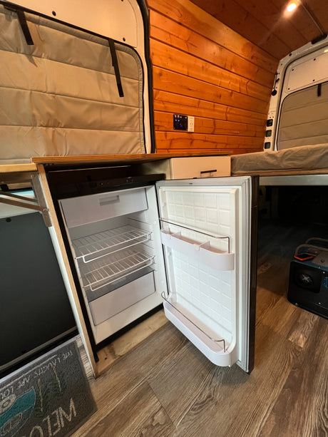 Open refrigerator in an rv rental van, showing wood paneling, controls, and a portable power station on a laminate floor.