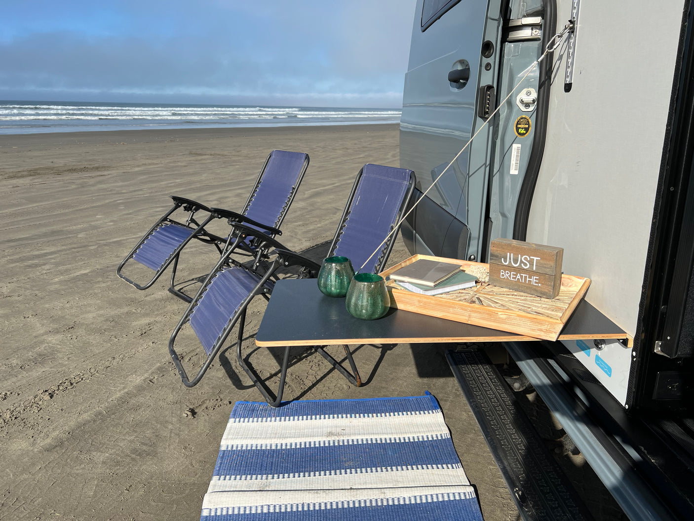 Peaceful RV rental beach day. A gray van with an attached table, two blue chairs, and a mat facing calming ocean waves.