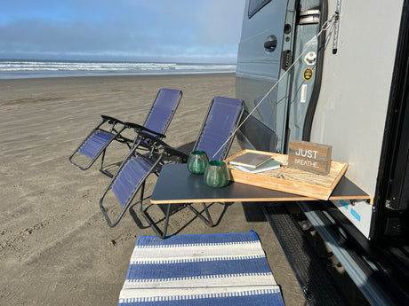 Peaceful RV rental beach day. A gray van with an attached table, two blue chairs, and a mat facing calming ocean waves.