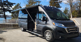 This grey Ram ProMaster rv rental is set up with its awning extended by a tranquil lake, ready for adventure.