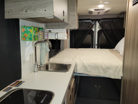 This rv rental features a compact kitchen with a sink and stove on the left, facing a cozy bed with white linens at the rear.