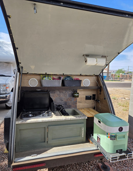 This organized teardrop RV rental's outdoor kitchen features a stove, sink, speakers, and a convenient pull-out cooler for camping.
