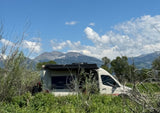 An RV rental camper van sits amidst green foliage, with snowy mountains under a bright blue sky. Perfect for nature exploration.