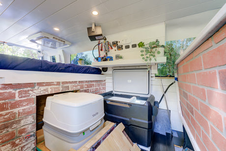 Compact RV rental interior: elevated bed, portable toilet, fridge, and built-in amenities in a converted van.