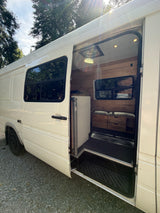 Open side door of a white camper van showing a custom wood interior, bench seat, and drawers. A perfect RV rental choice!