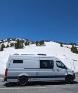 This white converted van, available for RV rental, is parked on a snowy mountain road under a clear blue sky.