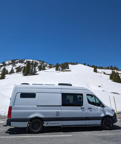 This white converted van, available for RV rental, is parked on a snowy mountain road under a clear blue sky.