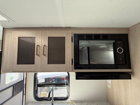 Inside an RV rental kitchen: light wood cabinets, Greystone microwave, range hood, sink, and window view.