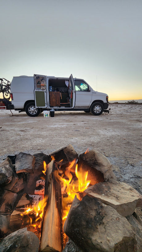 RV rental ready for adventure: A white camper van with an open door, a cozy campfire, and a dog in a desert sunset.