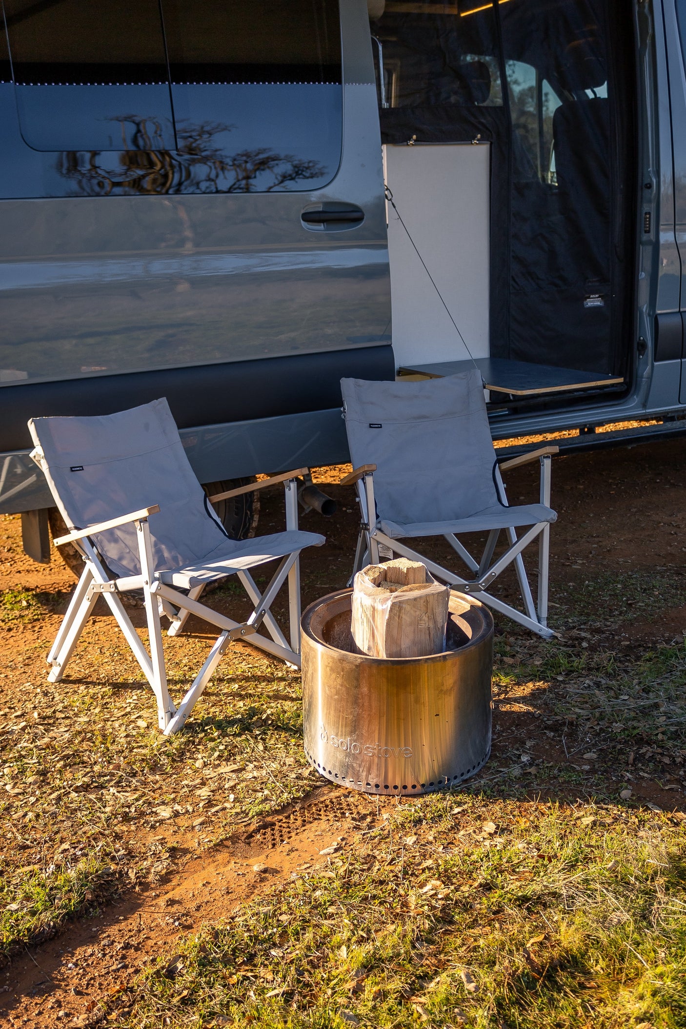 RV rental setup: Two camping chairs await a cozy evening around a Solo Stove fire pit beside a parked camper van.