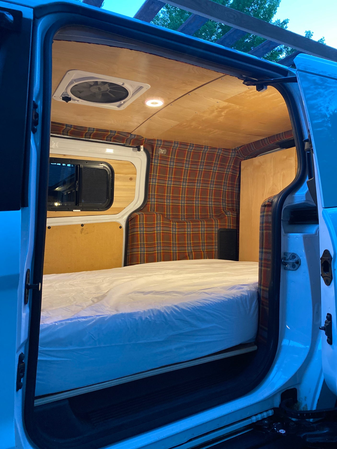 Cozy RV rental interior: bed, plaid walls, wood ceiling, fan, and window visible from the open door.