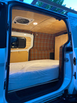 Cozy RV rental interior: bed, plaid walls, wood ceiling, fan, and window visible from the open door.
