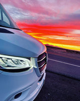 White RV rental van's headlight glows against a vibrant red, orange, and pink sunset sky over distant mountains.