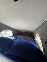 RV rental pop-top interior: a cozy bed with blue bedding and a striped blanket, illuminated by side windows.