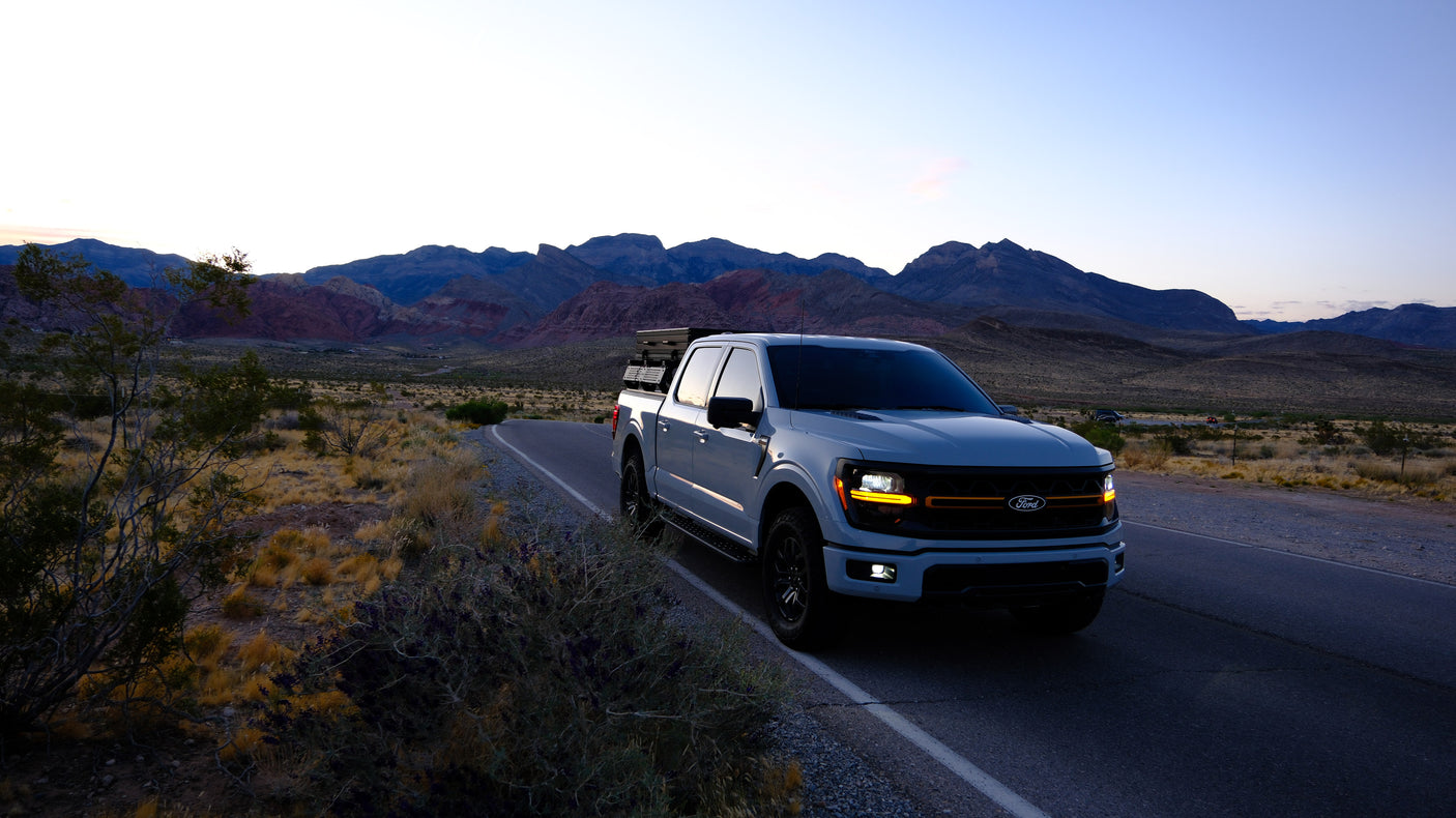 White Ford F-150 pickup truck with gear, parked roadside in a stunning desert mountain landscape at dusk. Great for your rv rental adventure.