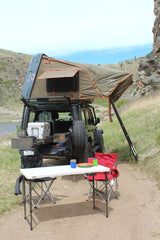 An overland Jeep with a deployed rooftop tent and camp setup is parked by a lake. Ideal for adventure travel as an RV rental alternative.