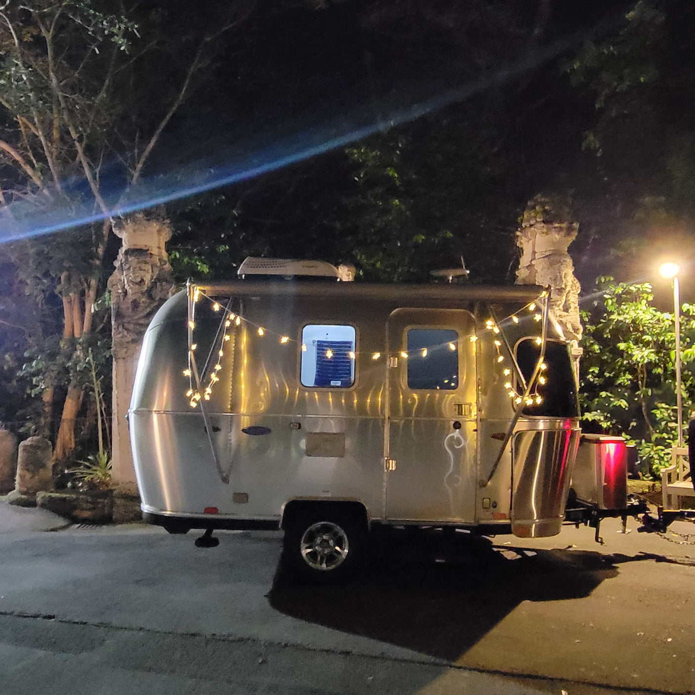 A shiny Airstream RV rental, decorated with star lights, parked at night under a bright lamp near trees.