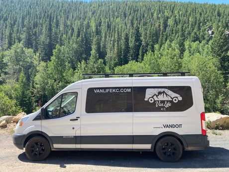 White camper van with "VANLIFEKC" branding, ideal for an **rv rental** adventure, parked roadside against a dense forest.