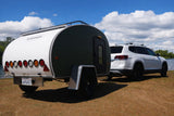 An RV rental adventure: White SUV tows a "CALIFORNIA TEARDROP" trailer by a scenic lake, perfect for your next road trip.