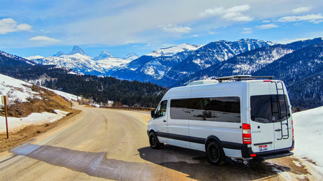 White Sprinter RV rental parked on a snowy mountain road, overlooking majestic, snow-capped peaks.