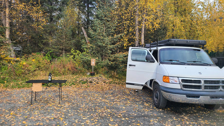 White RV rental camper van parked on autumn leaves by trees, with a table. Overnight parking allowed for fall adventures.
