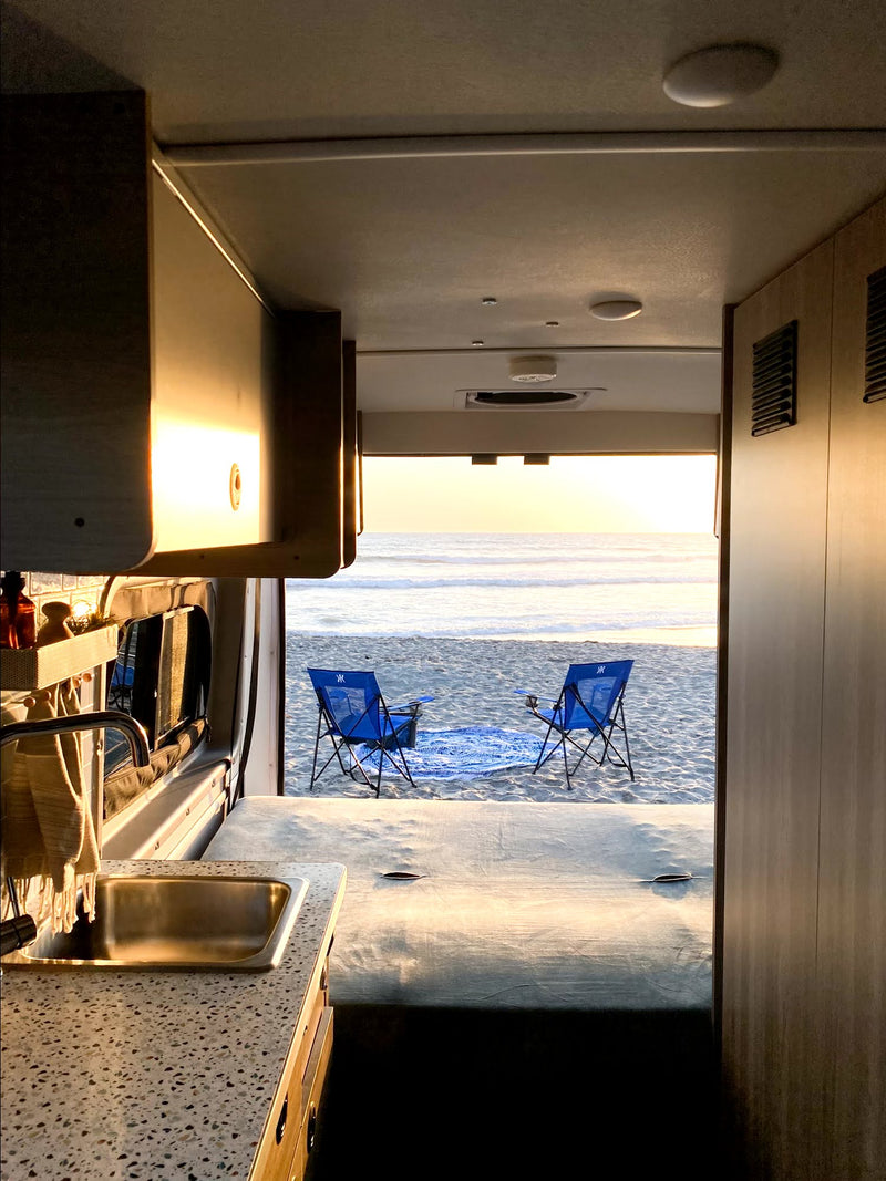 RV rental interior view: sink/counter, open back revealing a beach at sunset with two blue chairs by the ocean. — photo 8