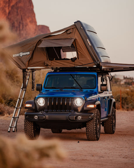A blue Jeep Wrangler with a Desert Armor rooftop tent deployed, ladder extended. Great for an RV rental off-road camping adventure.