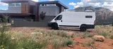 Explore with an RV rental! This white camper van is parked by a modern home, set against majestic red rock mountains.